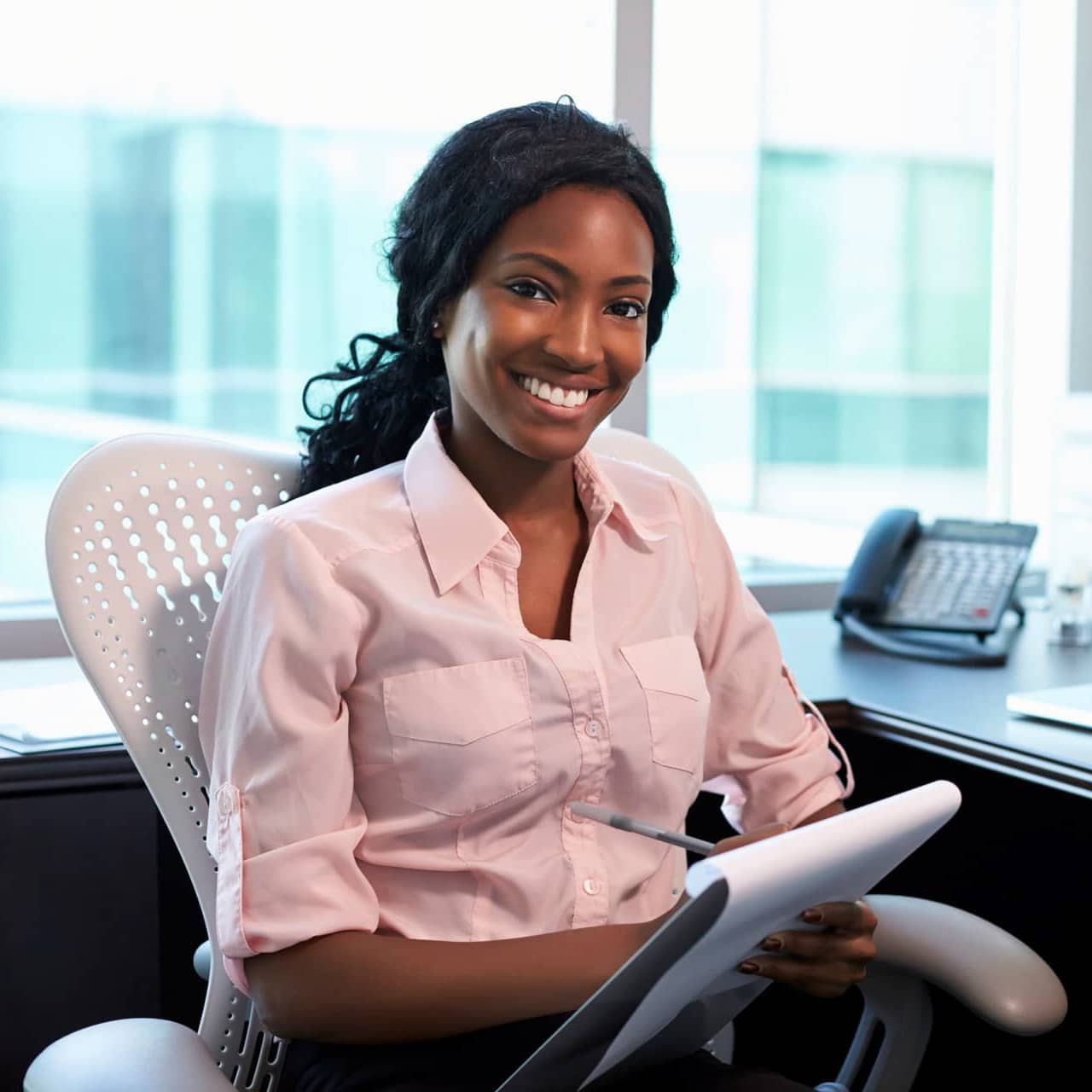 A business woman smiling while looking at you at camera in office with notepad in a pink blouse.